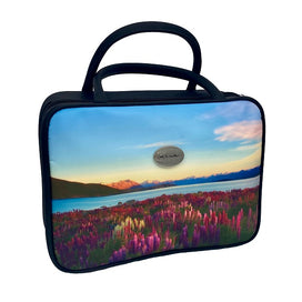 A rectangular vanity/cosmetic case with 2 black handles. The front and back of the cosmetic case display a beautiful scene of pink and purple lupins, with New Zealand’s spectacular mountain ranges in the background, under a blue sky with wispy clouds. One side of the cosmetic case also features a silver badge with the AA Traveller logo. The sides are black, with a black zip around 3 sides. 